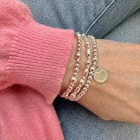 Close-up of a wrist wearing multiple silver beaded bracelets with a heart disc charm, against a pink sleeve and denim background.