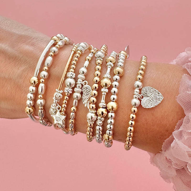 Close up of a wrist wearing several mixed metal (gold/silver) beaded stacking bracelets with charms, against a pink background
