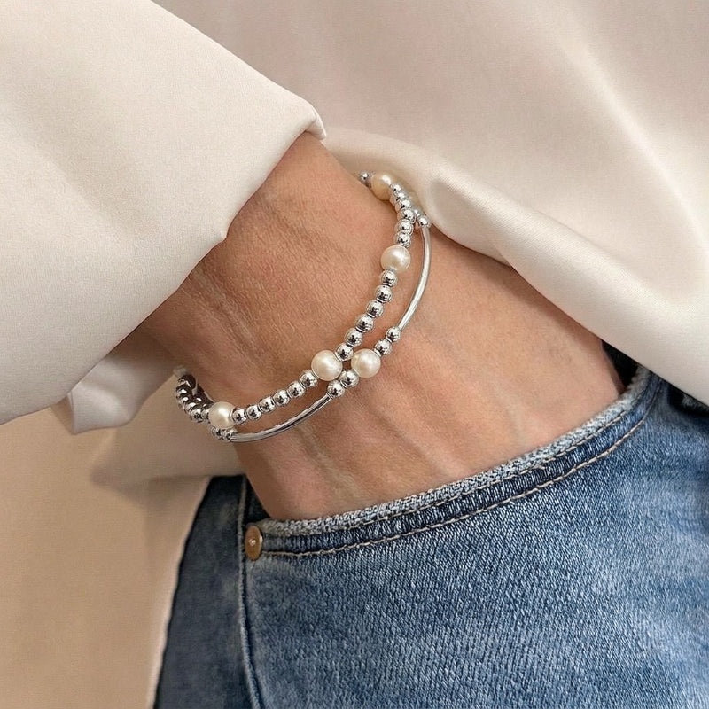 A close-up of a person's wrist wearing two sterling silver beaded bracelets featuring freshwater pearl beads, styled with a white blouse and blue jeans.