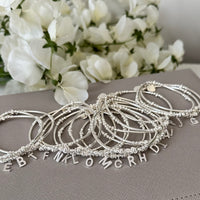 A selection of sterling silver beaded bracelets all featuring different sparkly initial charms, resting on a grey surface with white flowers in the background