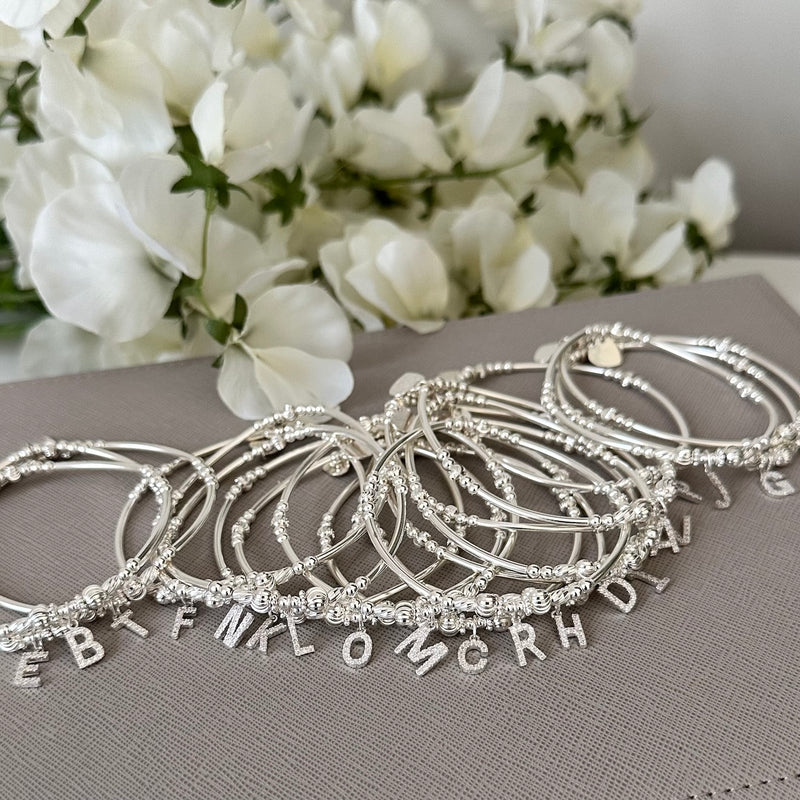A selection of sterling silver beaded bracelets all featuring different sparkly initial charms, resting on a grey surface with white flowers in the background