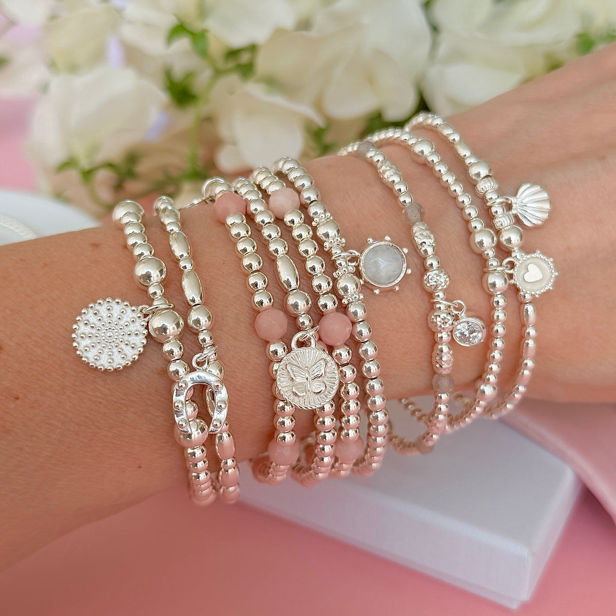 A close-up of a person's wrist wearing multiple beaded stacking bracelets in silver and soft pink tones, with charms including a shell, heart, flower, and round stone, against a pink and white background with blurred white flowers.