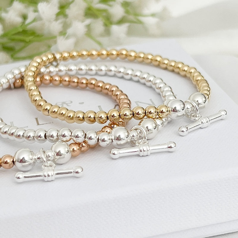 Set of three beaded bracelets in gold, rose gold, and silver with silver t-bar charms on a white surface with a blurred floral background.