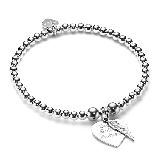 A silver stacking bracelet with a silver heart-shaped charm engraved with 'Dream believe achieve' with a tiny angel wing charm on a white background