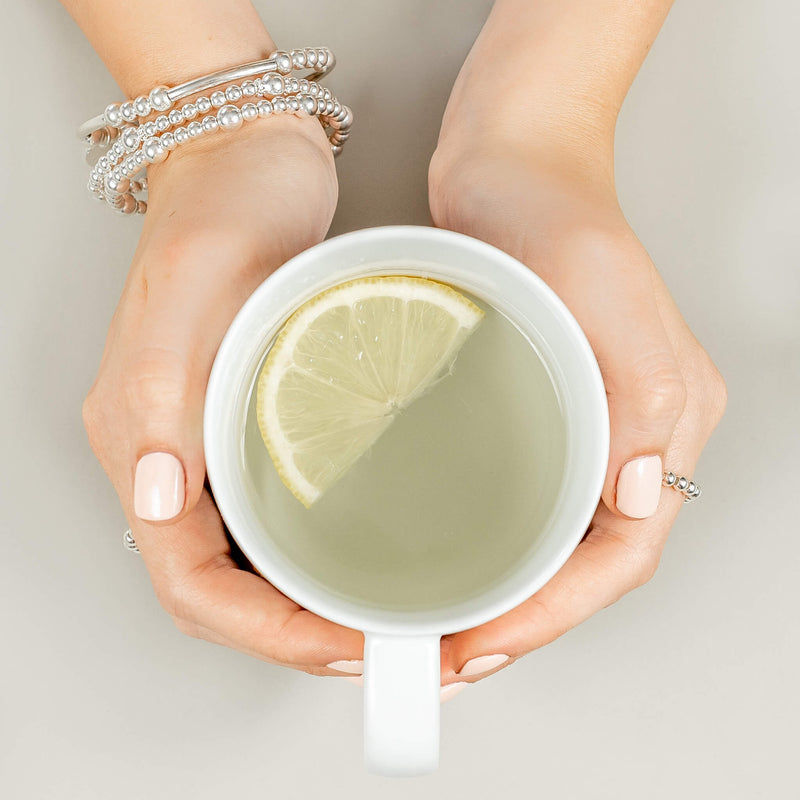 A set of three sterling silver stacking bracelets modelled on a wrist holding a mug of lemon water