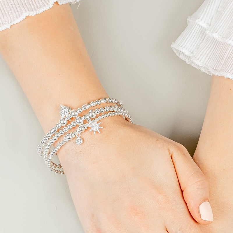 Close-up of a wrist wearing three silver bracelets with a sparkly starburst and round charm with a faceted cut heart charm, against a neutral background