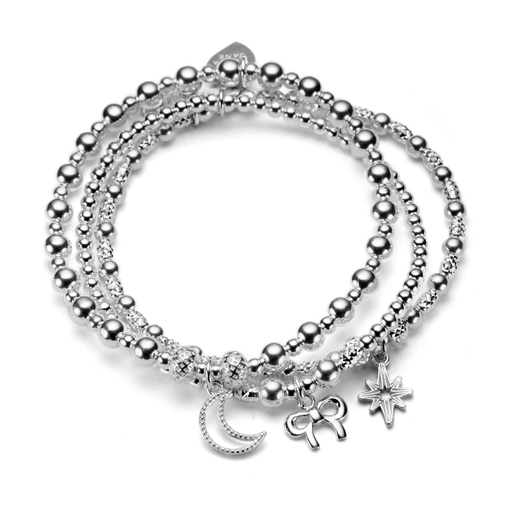 A trio of silver beaded stacking bracelets, one with a moon, one with a bow and one with a guiding star charm, against a white background.