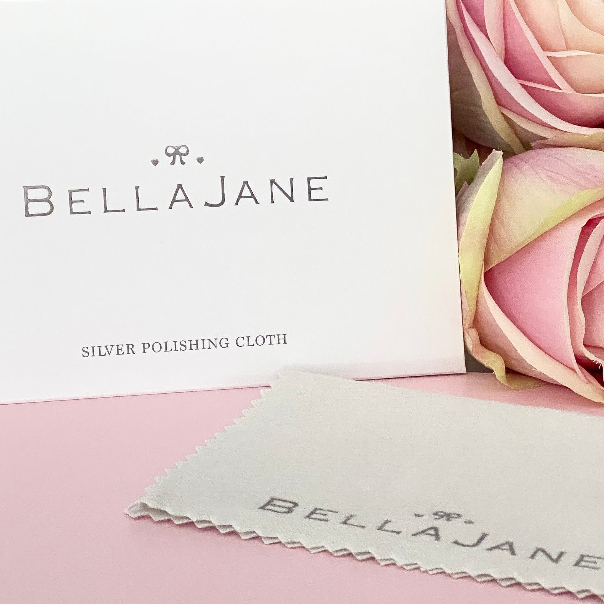A silver polishing cloth with the Bella Jane brand logo displayed beside some pink roses.
