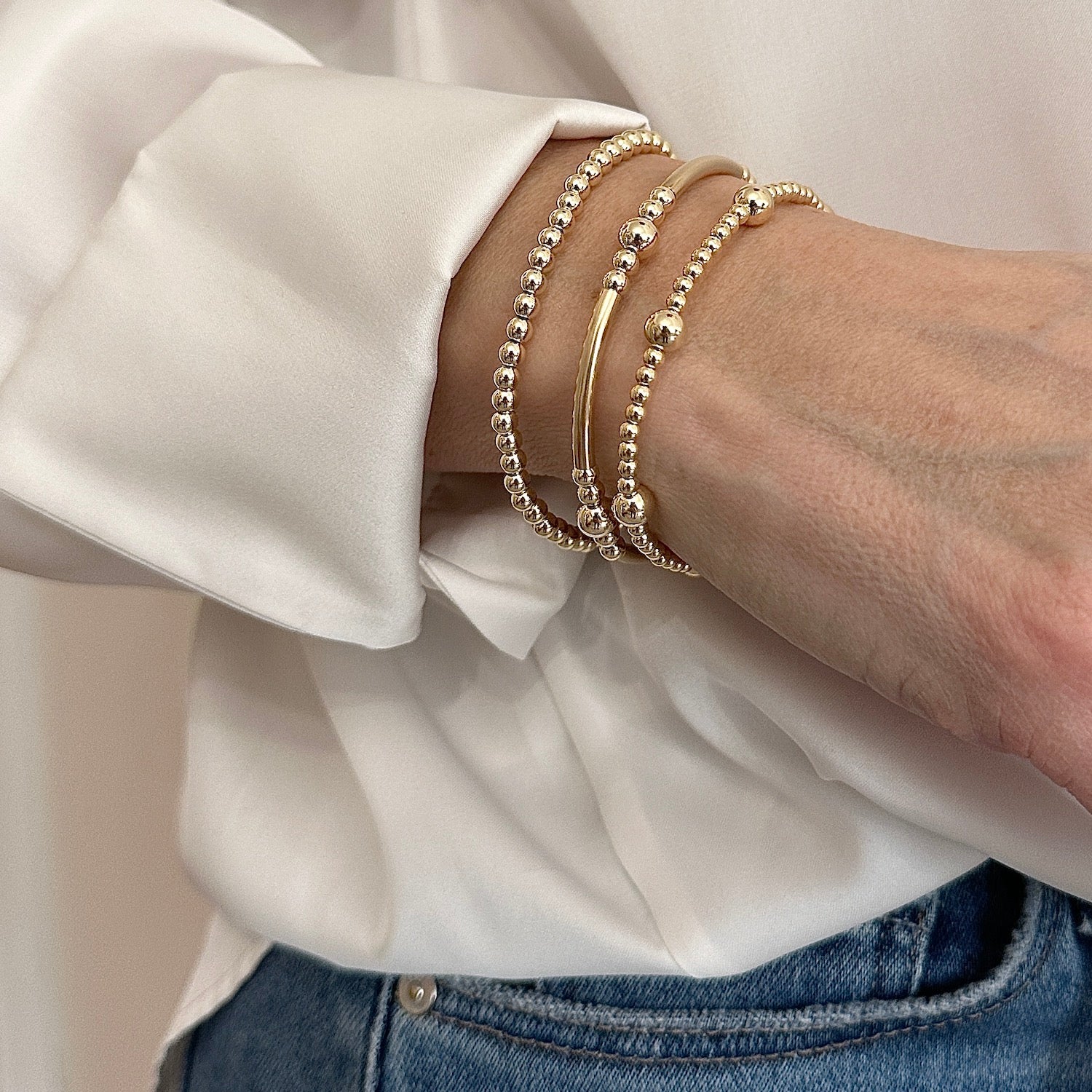 A model showing her wrist wearing a stack of 3 gold-filled beaded stacking bracelets. The model is wearing jeans and an ivory coloured silk shirt