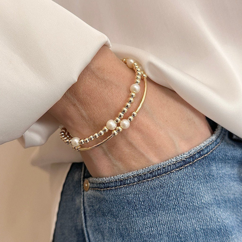 Two gold beaded bracelets with pearls on a wrist, wearing a white shirt and blue jeans.