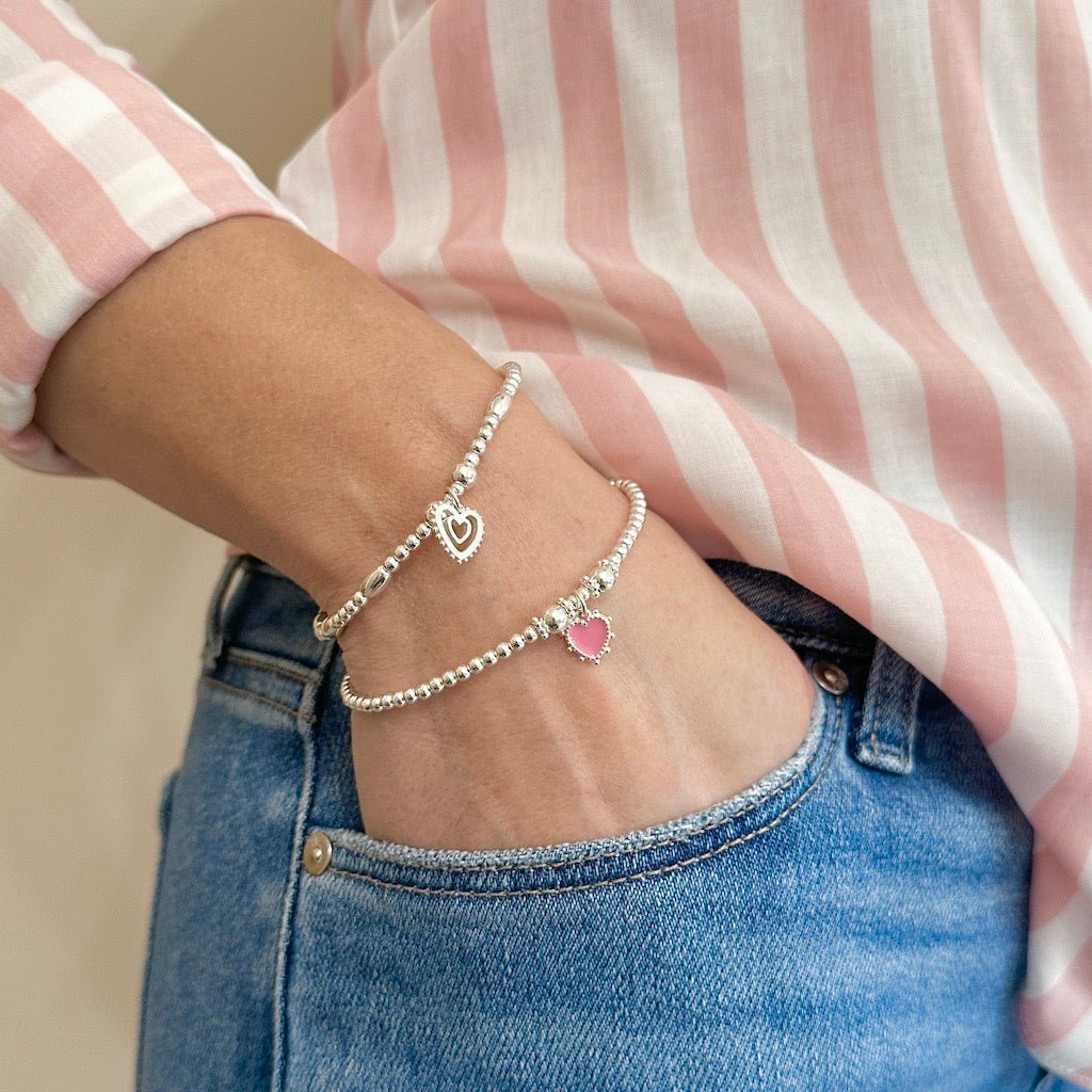 Person wearing two silver stacking bracelets with heart charms with a pink and white striped shirt and jeans