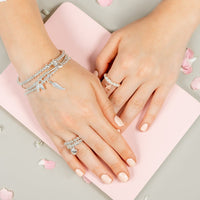 Close-up of hands wearing silver stacking bracelets with angel wings, heart and feather charms, resting on a pink notebook with scattered rose petals.