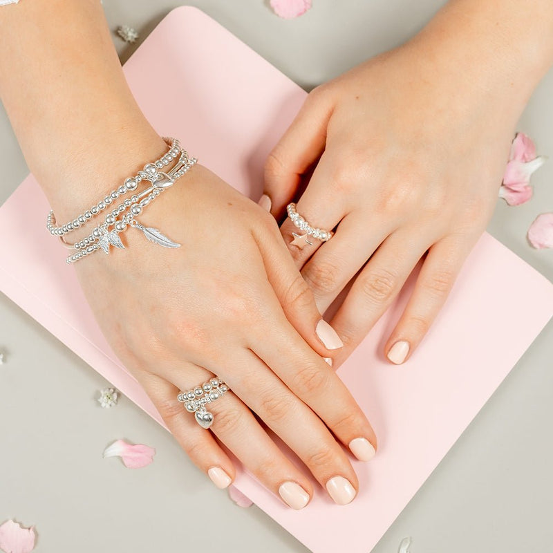 Close-up of hands wearing silver stacking bracelets with angel wings, heart and feather charms, resting on a pink notebook with scattered rose petals.