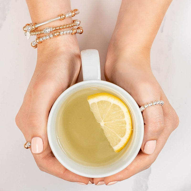Hands holding a cup of tea with a lemon slice, wearing silver and rose gold beaded stacking bracelets and rings on a light background