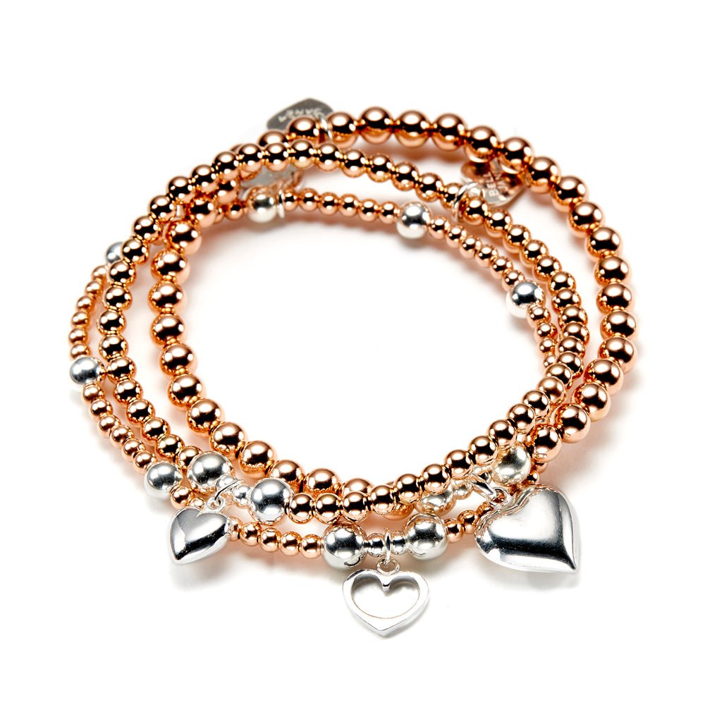 A stack of three rose gold and silver beaded bracelets, featuring three different sterling silver heart charms, displayed on a white background