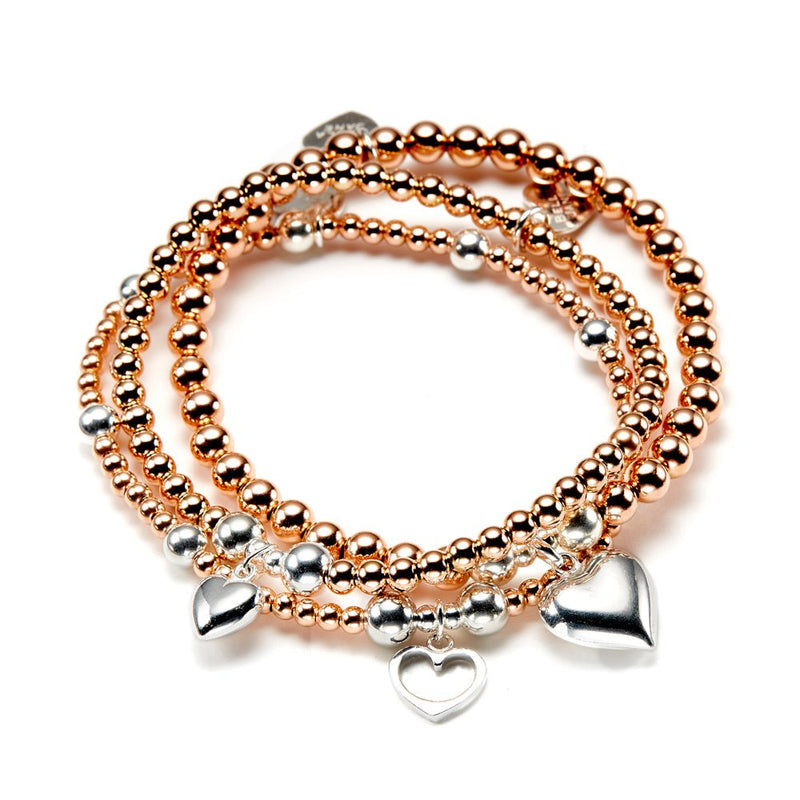 A stack of three rose gold and silver beaded bracelets, featuring three different sterling silver heart charms, displayed on a white background